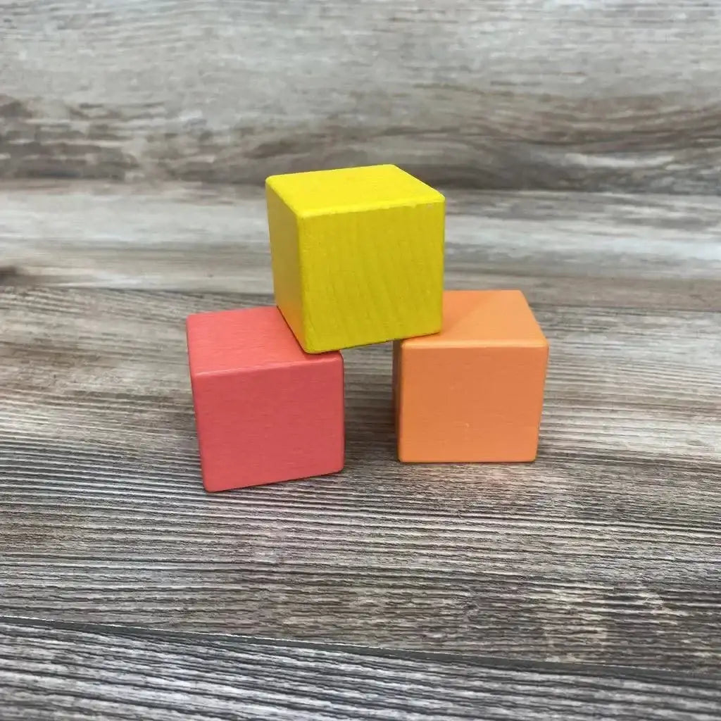 Three-quarter view of Lovevery 3-piece First Blocks showing yellow, coral, and orange painted wooden cubes stacked on a wood-grain surface