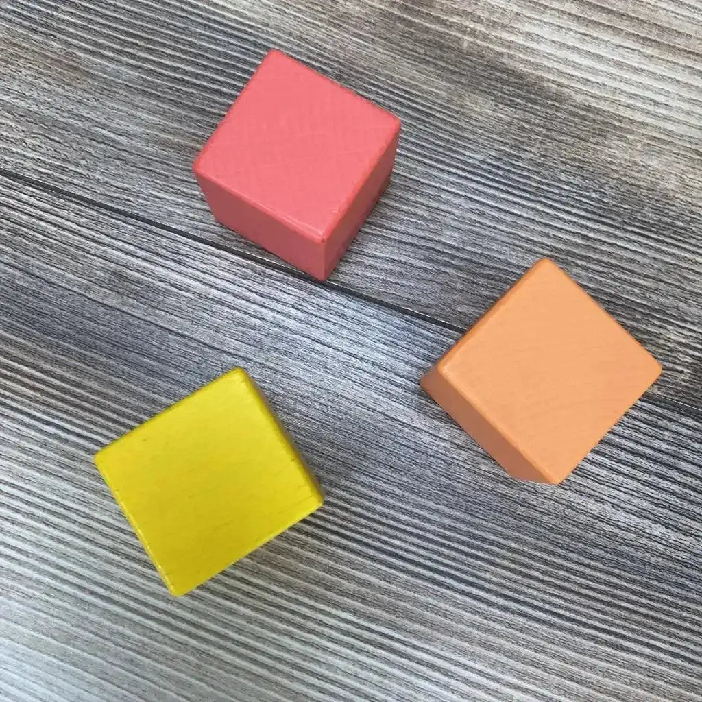 Angled top view of three Lovevery wooden blocks (yellow, orange, coral) on gray woodgrain floor