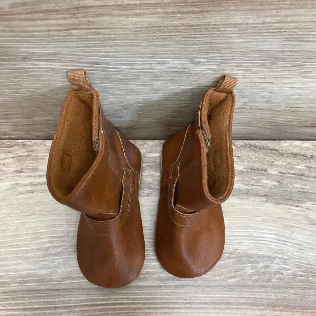 Top-down view of brown faux-leather tall baby booties showcasing buckled strap, pull tabs, and soft lining on light wood background