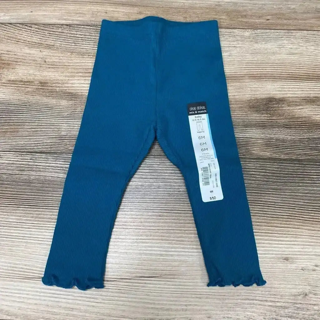 Flat lay of teal Okie Dokie ribbed baby leggings (6M) showing scalloped ankle hems and attached size tag on wood background