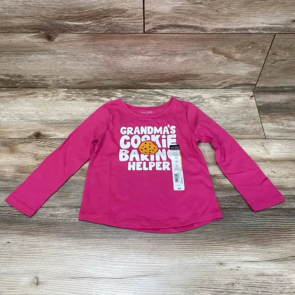 Flat lay of pink toddler long-sleeve shirt size 3T with text GRANDMA'S COOKIE BAKIN' HELPER and cookie graphic on wood floor
