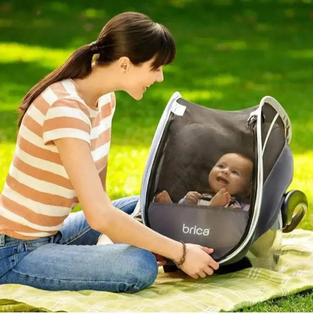 Woman sitting on a picnic blanket smiling at a baby inside a grey infant car seat with a mesh comfort canopy in a sunny park