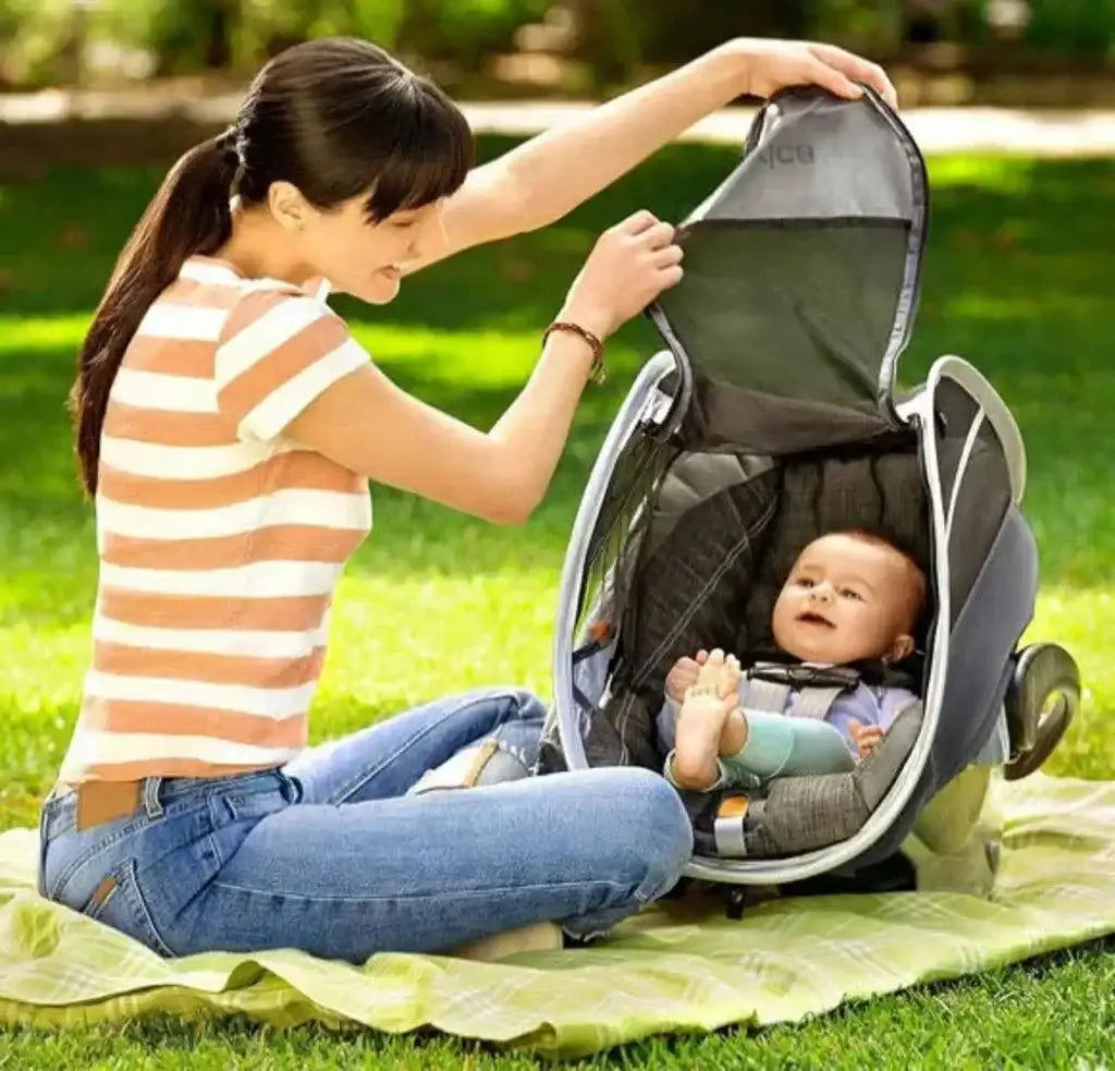 Woman placing grey Brica infant car seat canopy over a baby in a rear-facing car seat on a picnic blanket in a park