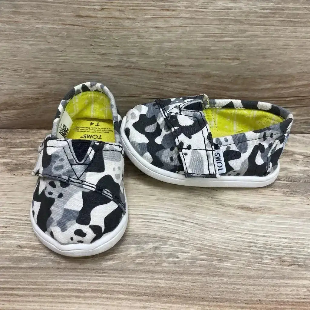Front and side view of TOMS slip-on shoes with black, grey and white camo, Velcro strap, yellow lining on wood background