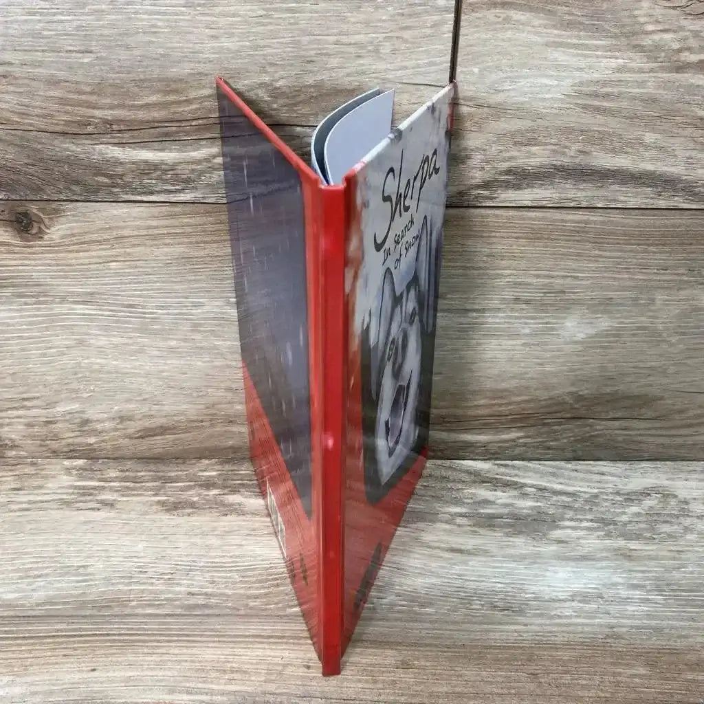 Angled view of used Sherpa hardcover book showing red spine and dog illustration on front, wood background
