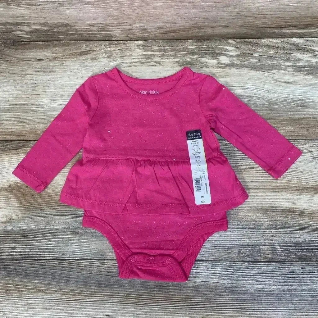 Front view of pink Okie Dokie skirted bodysuit in size 3M with long sleeves and layered skirt on wood-grain background