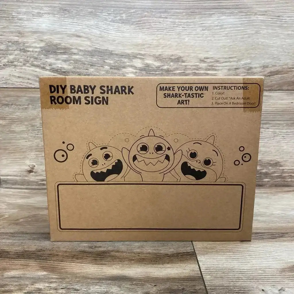 Front of Baby Shark DIY room sign box showing three cartoon sharks and a blank nameplate on a wood plank background