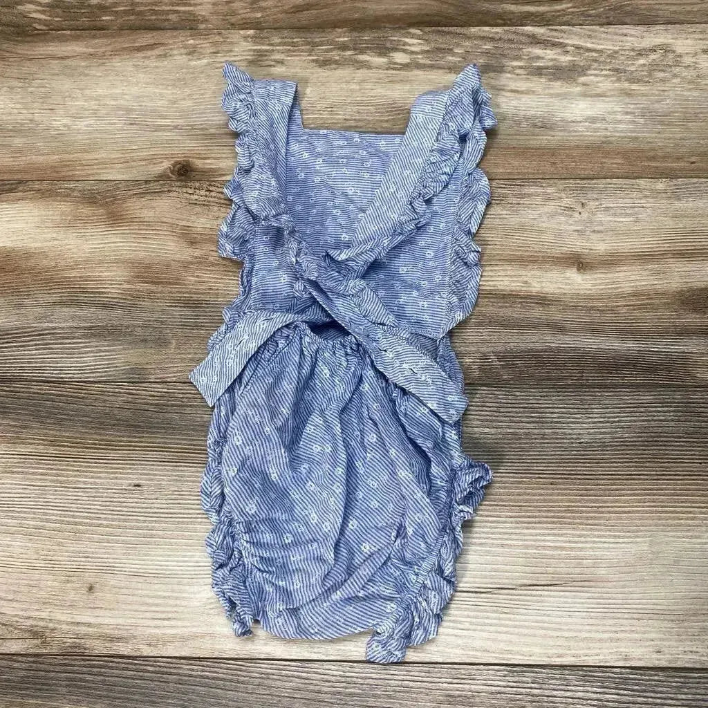 Flat lay of blue numi floral romper (3T) with crossed ruffle straps, waist tie and ruffled elastic legs on wood background
