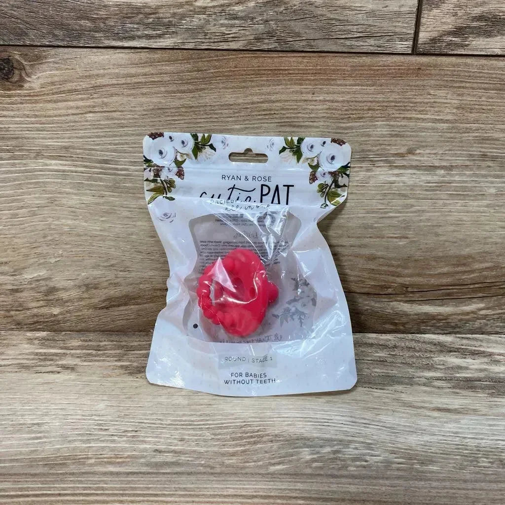 Front view of Ryan & Rose Cutie PAT round pacifier and teether in pink inside clear retail pouch on wood background