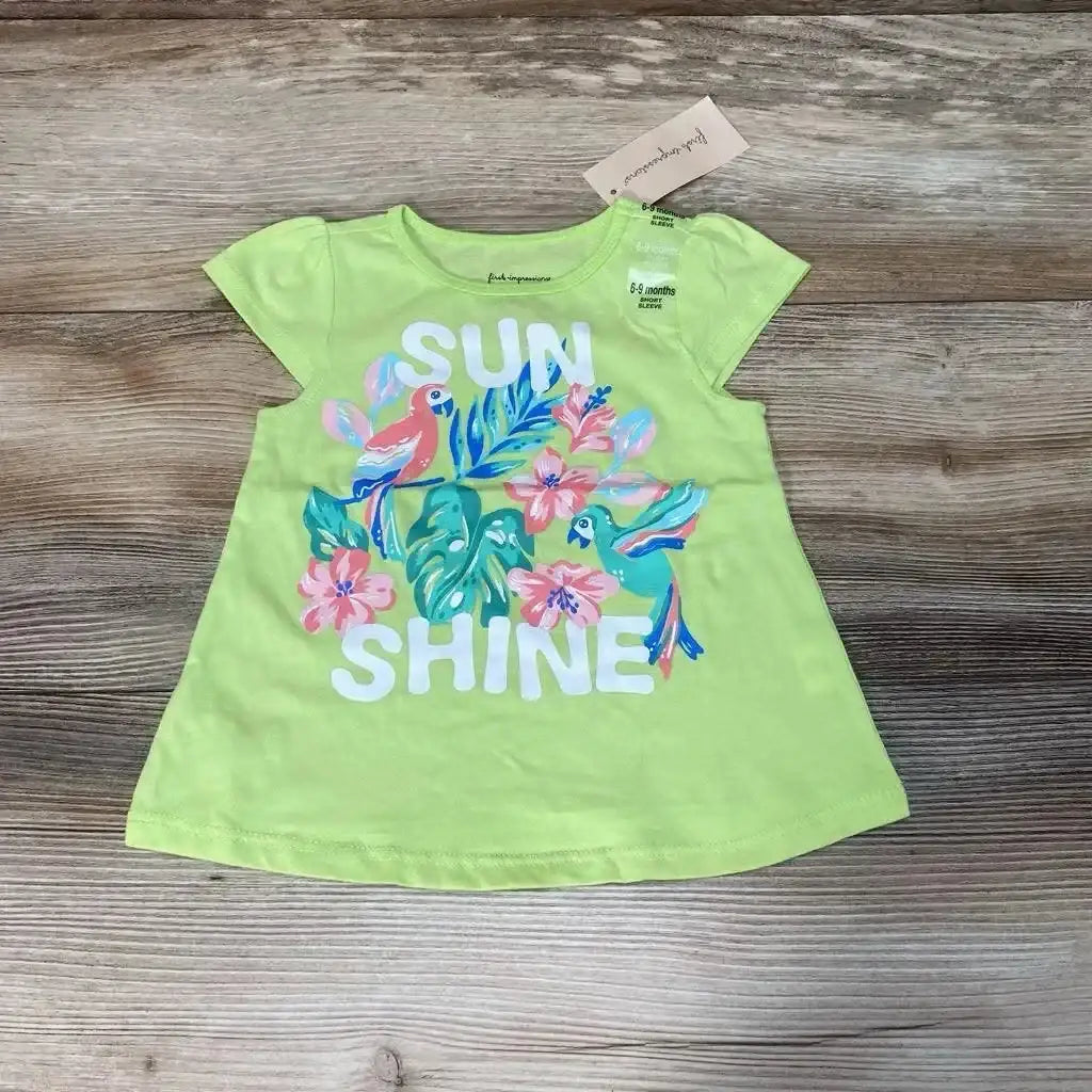 Flat lay of green First Impressions Sun Shine baby shirt (6–9 months) showing tropical parrot and flower graphic with large 'SUN SHINE' text on a light wood background