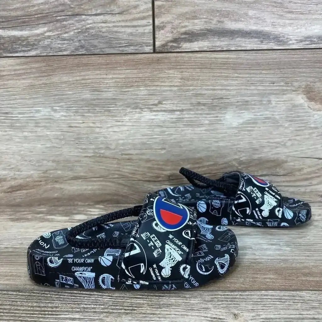 Side view of black Champion Super Slide toddler sandals (size 8C) showcasing white doodle sports print and Champion logo on wooden floor background