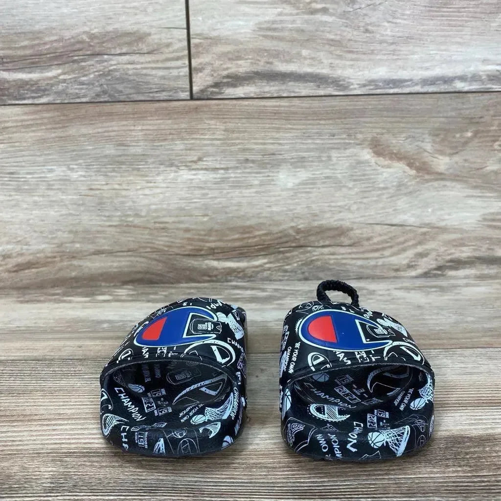 Front view of black Champion toddler slide sandals showcasing doodle print and Champion logo on straps with wood floor background