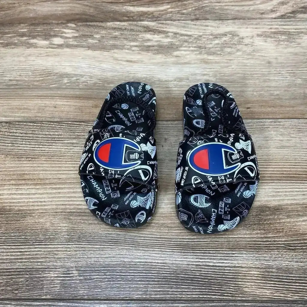 Top-down view of black Champion Super Slide doodle sandals size 8C showing printed doodle pattern and large Champion logo on wood plank background