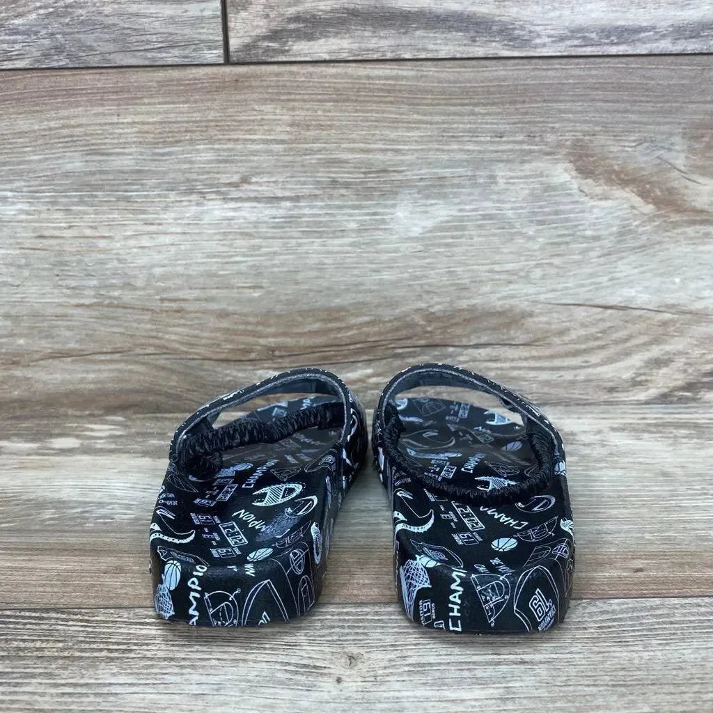 Rear view of black Champion Super Slide Doodle toddler sandals showing doodle print and heel straps on light wood floor