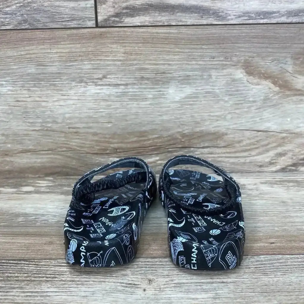 Rear view of black Champion Super Slide Doodle toddler sandals showing doodle print and heel straps on light wood floor