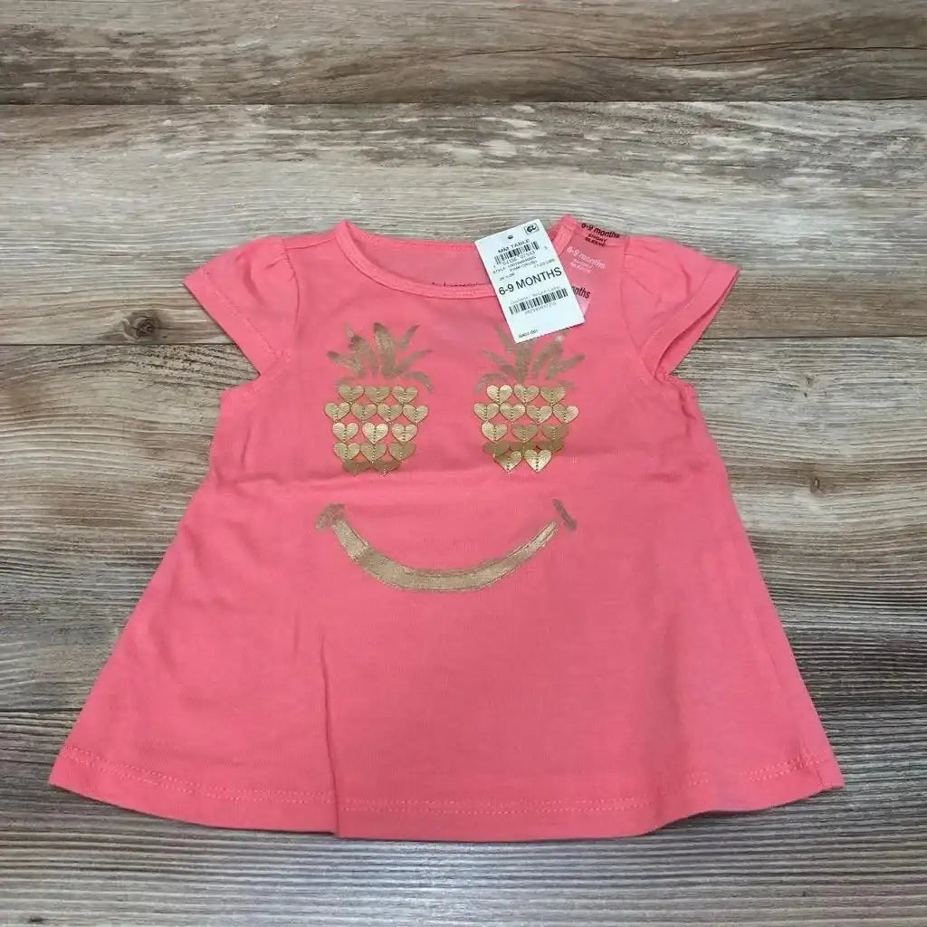 Front view of pink 6–9 month baby shirt showcasing gold pineapple heart eyes and gold curved smile, flat on wooden surface