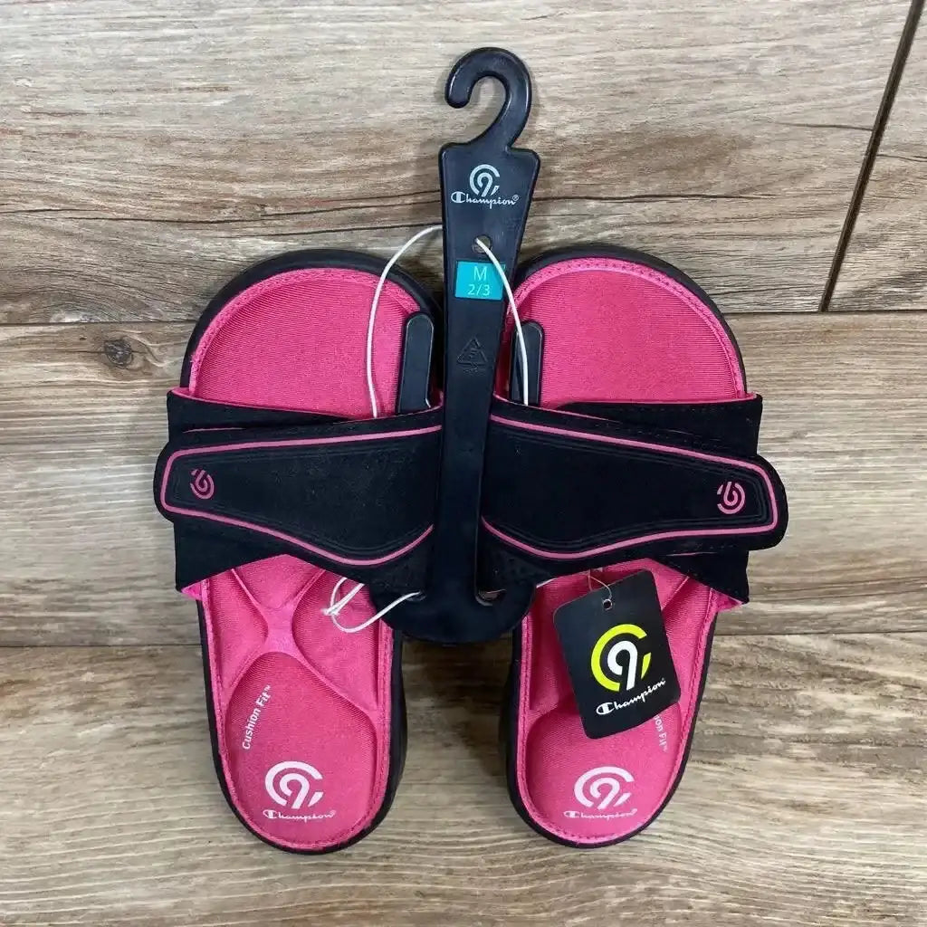 Overhead view of pink Champion memory-foam toddler slide sandals with black adjustable straps on wood background, size 2/3Y