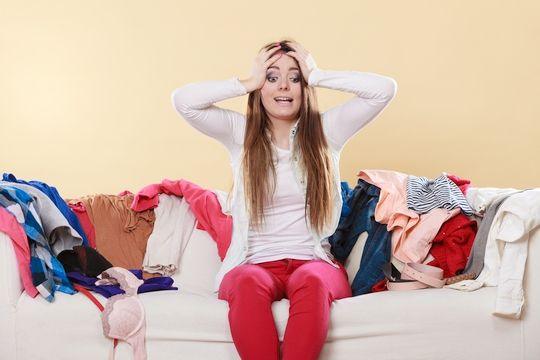 Woman sitting on a sofa surrounded by piles of clothes, looking overwhelmed while sorting laundry for spring cleaning