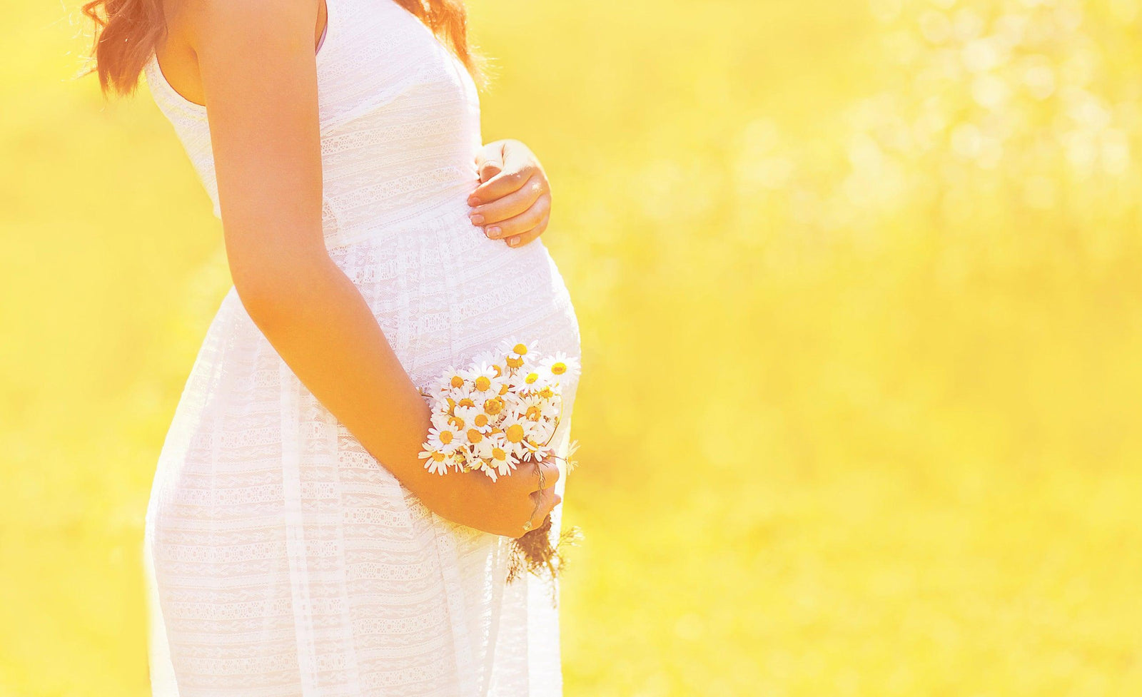 Pregnant woman in white sundress holding a small bouquet of daisies in a bright sunlit field