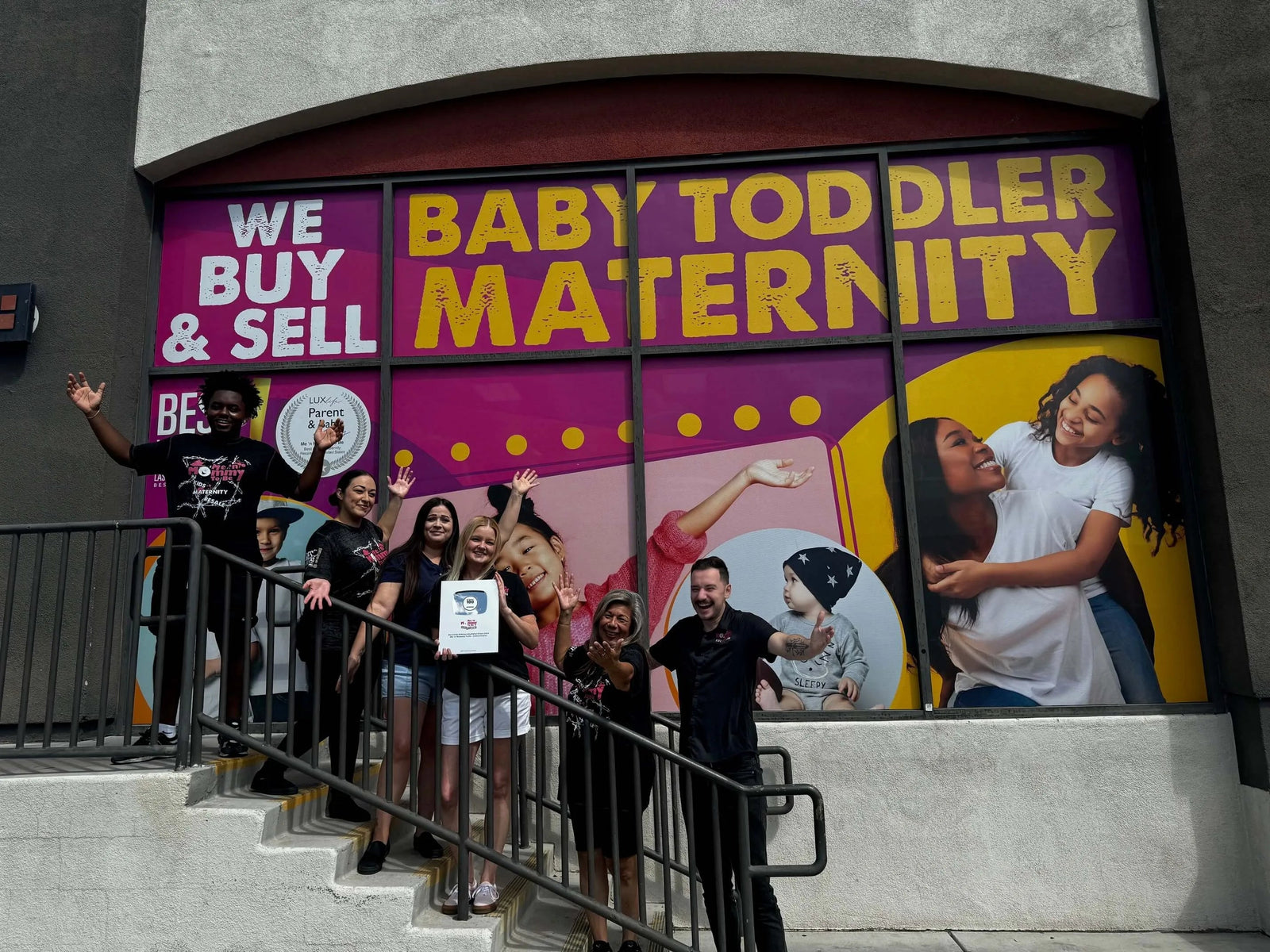 Store staff celebrating with award on storefront steps in front of large BABY TODDLER MATERNITY window mural