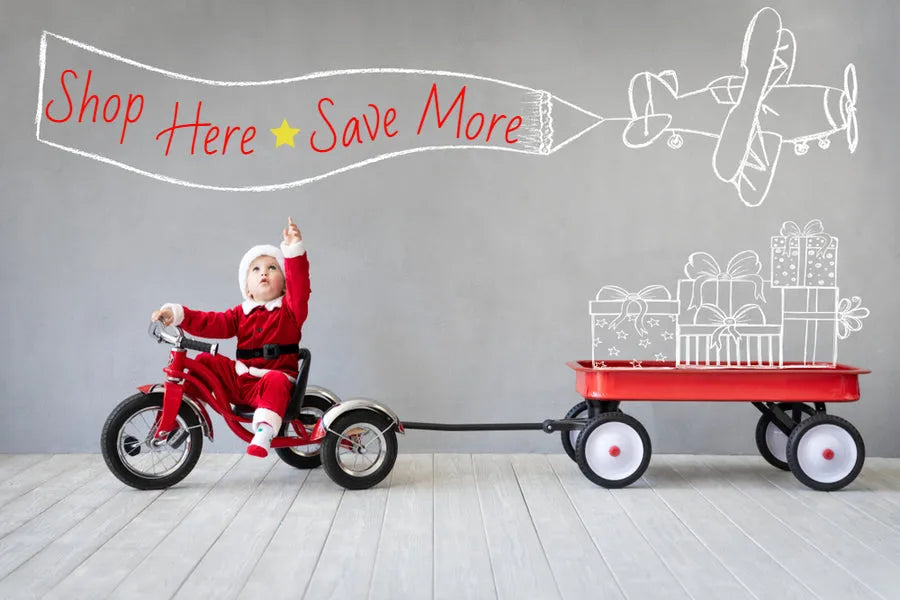 Baby in Santa suit on red tricycle towing a red wagon with drawn gifts against gray wall, banner text Shop Here Save More