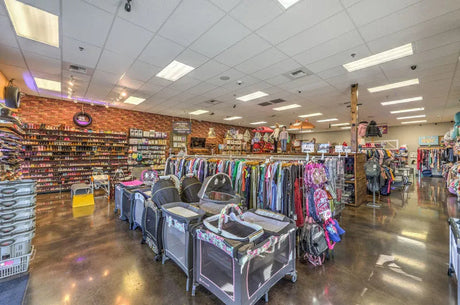 Resale store interior showing rows of children's clothing racks, several playpens in the foreground, and shoes displayed on the back wall