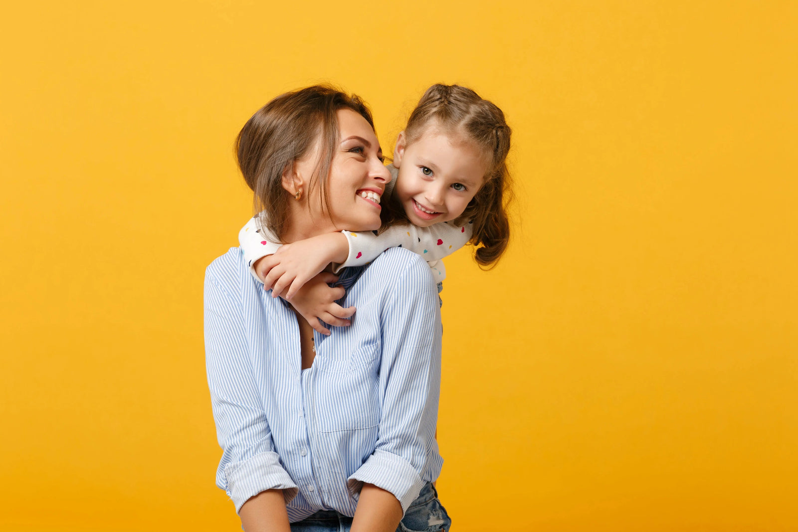 Mother carrying her smiling young daughter on her back as the child hugs her shoulders against a bright yellow background