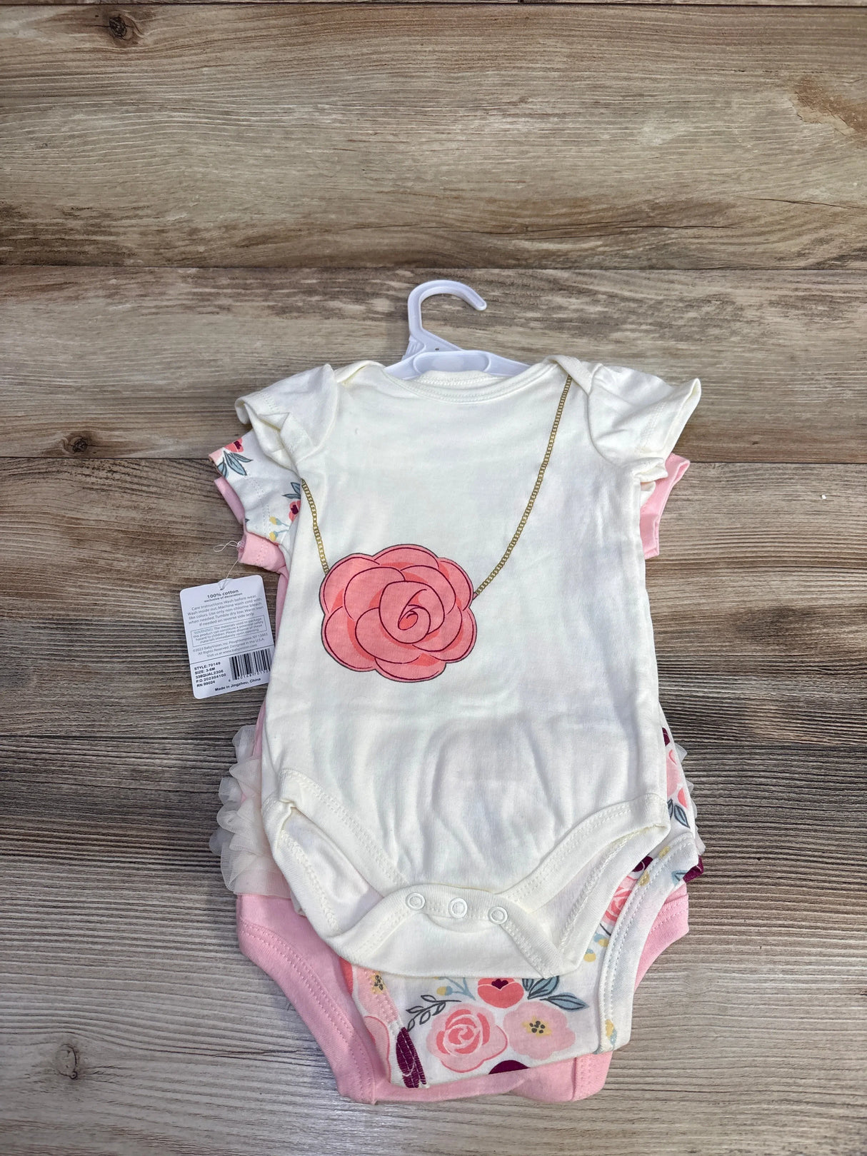 Top view of 3-pack baby girl bodysuits: white with pink rose necklace print, floral and pink, on wood background