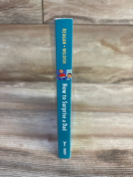 Side view of How to Surprise a Dad board book spine showing teal cover, author Reagan Wildish, and Knopf logo on wood background