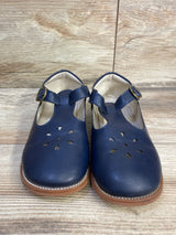 Front view of navy T-strap Mary Jane flats with flower cutouts and brass buckle on tan sole against wood floor background