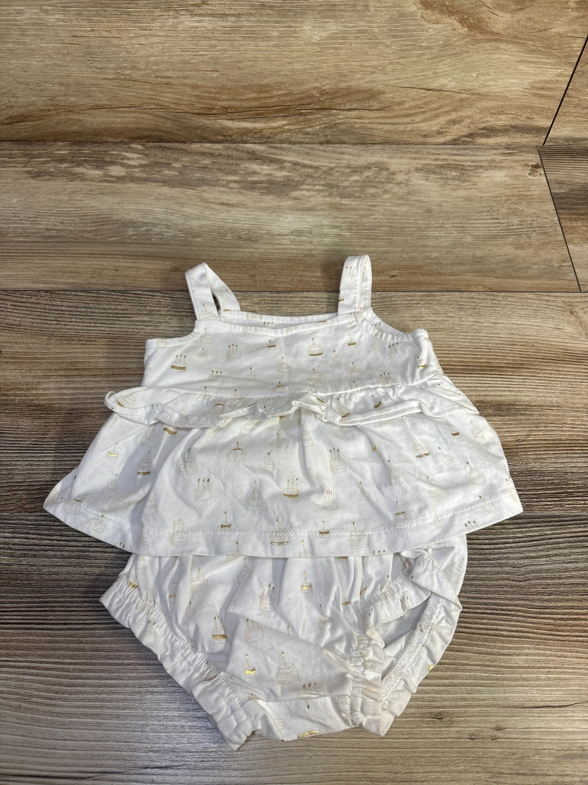 Top view of Baby Gap white 2pc Birthday Cake outfit ruffle tank and bloomers with gold cake print on wood floor