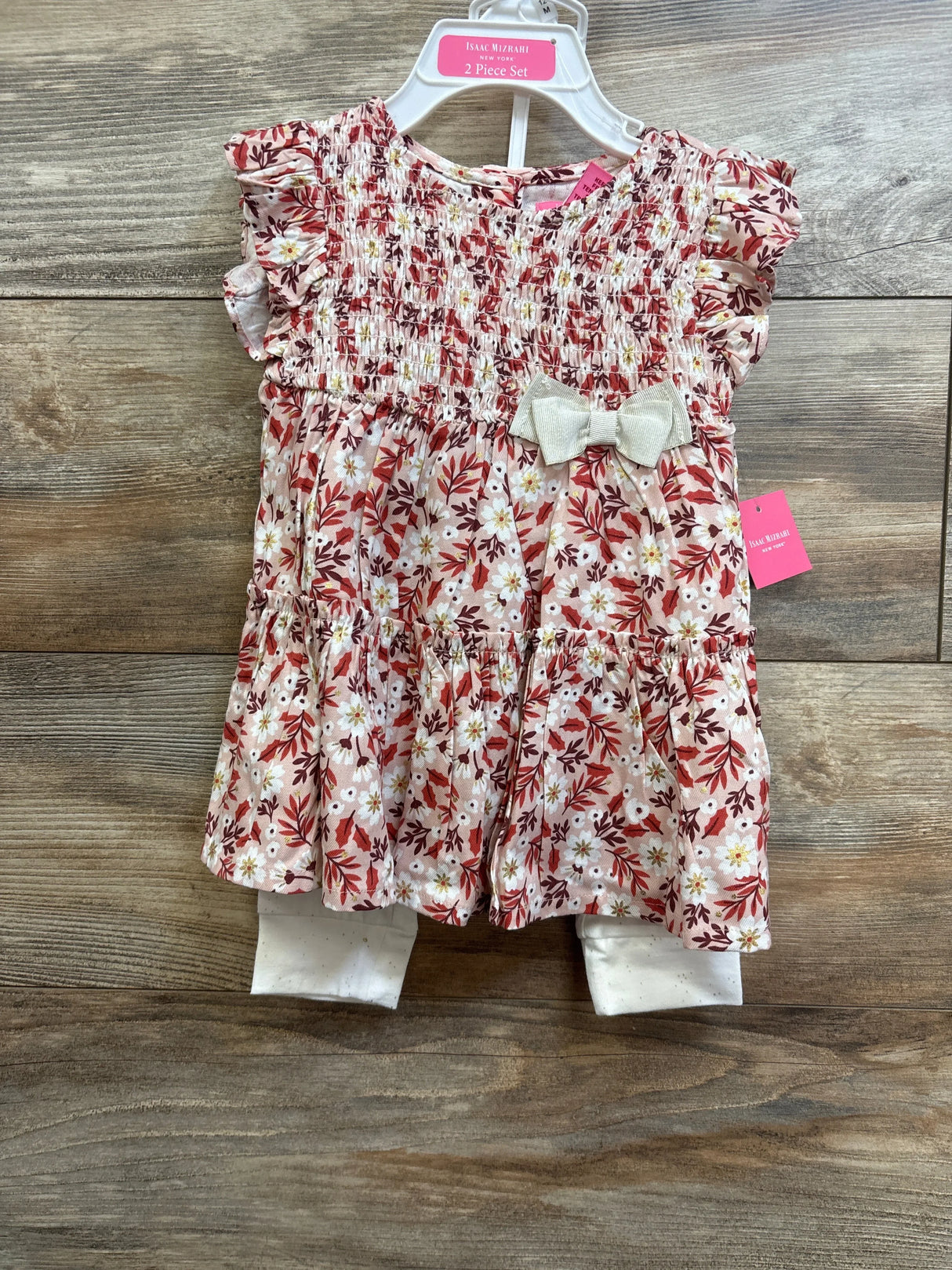 Front view of toddler 2-piece pink smocked floral dress and white leggings set showcasing smocked bodice, ruffle sleeves and ivory bow on wooden background