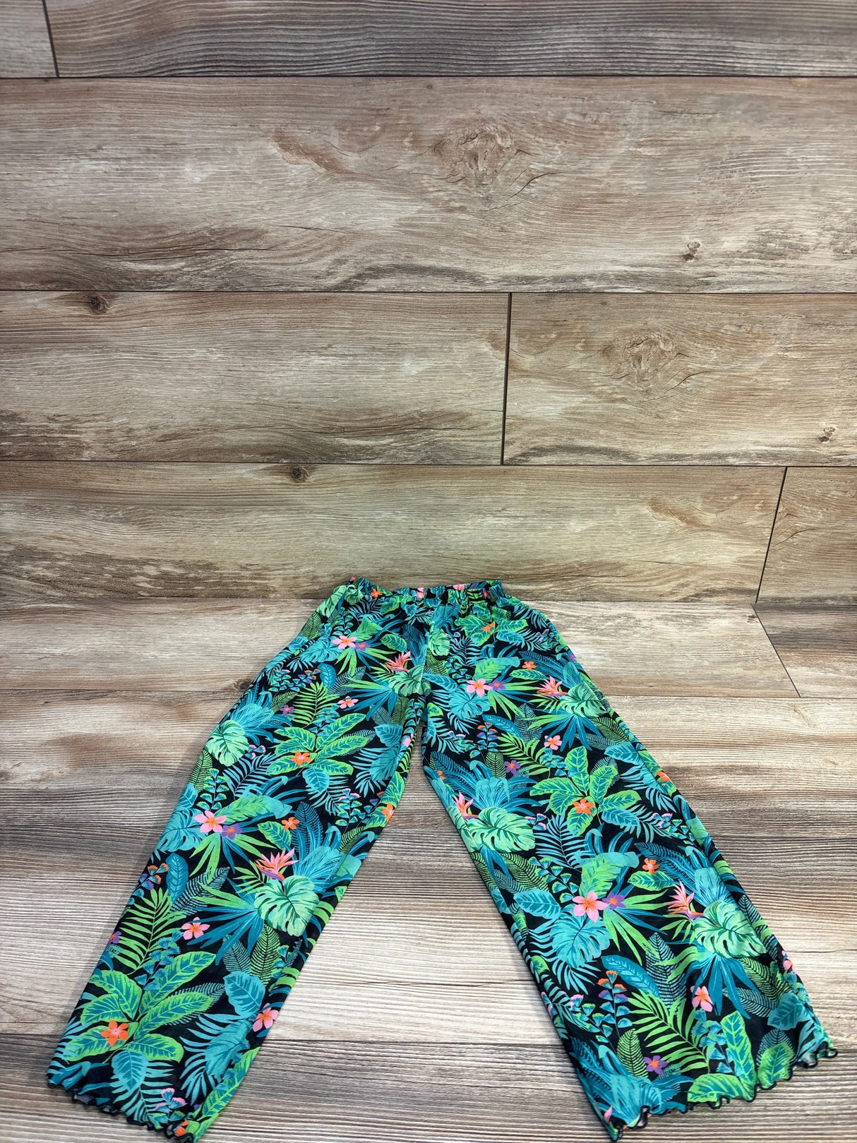 Flat lay of teal tropical print swim cover up pants showing elastic waistband and ruffled ankle hems on wood background