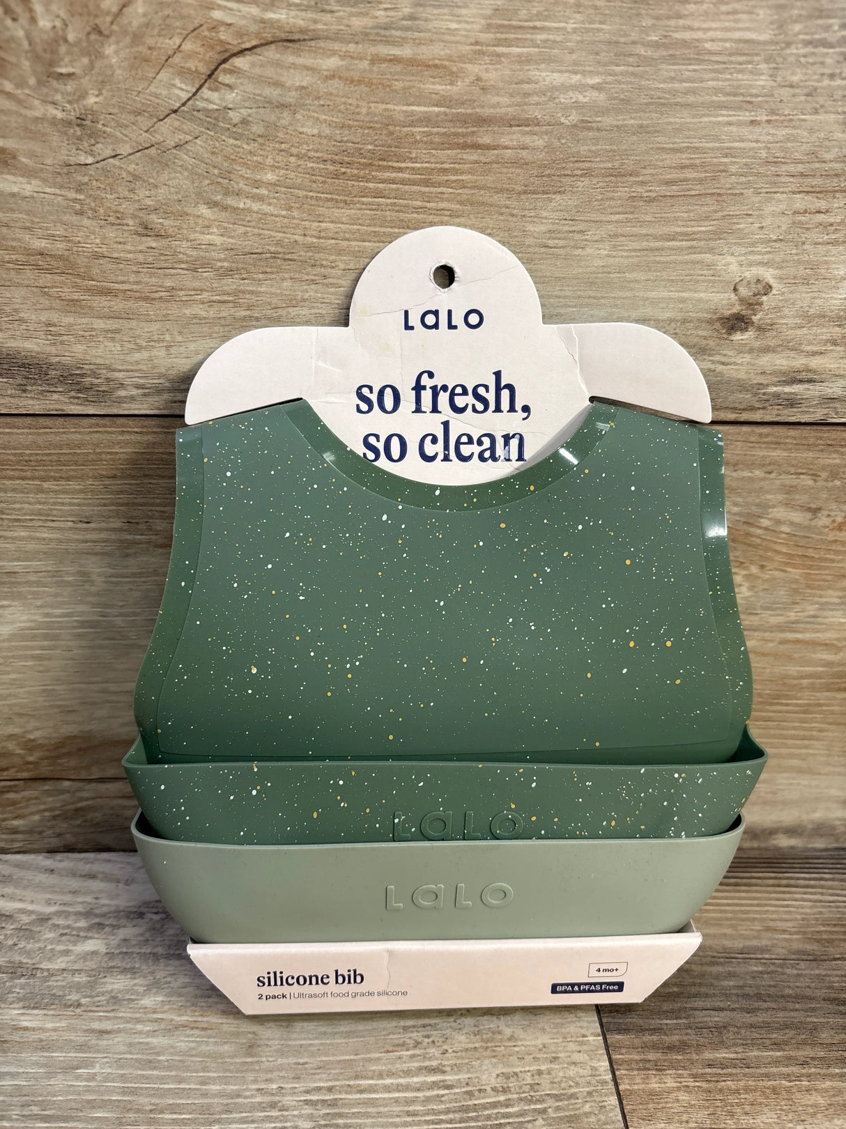 Front view of Lalo 2-pack silicone bibs in sage showing speckled top bib and deep crumb-catching pocket on wood background