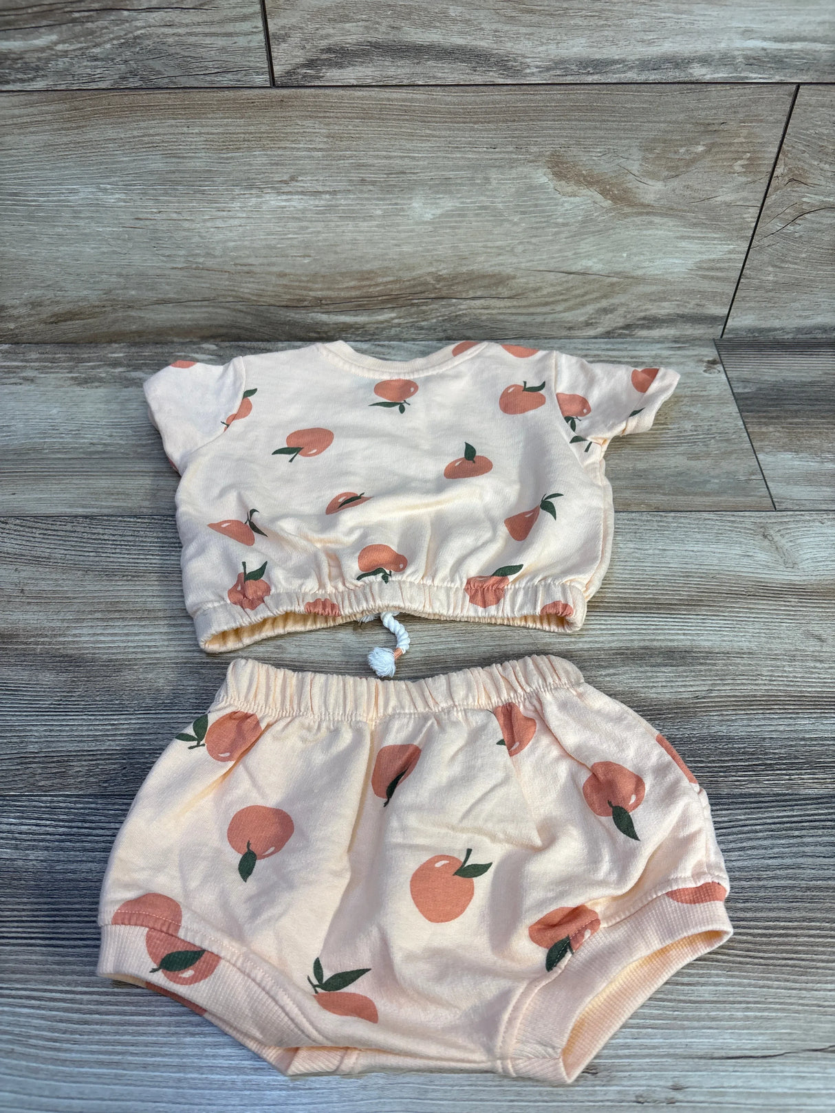 Top-down view of Jessica Simpson 2pc shirt and shorts set, size 3-6M, peach with peach print and elastic waists on wood