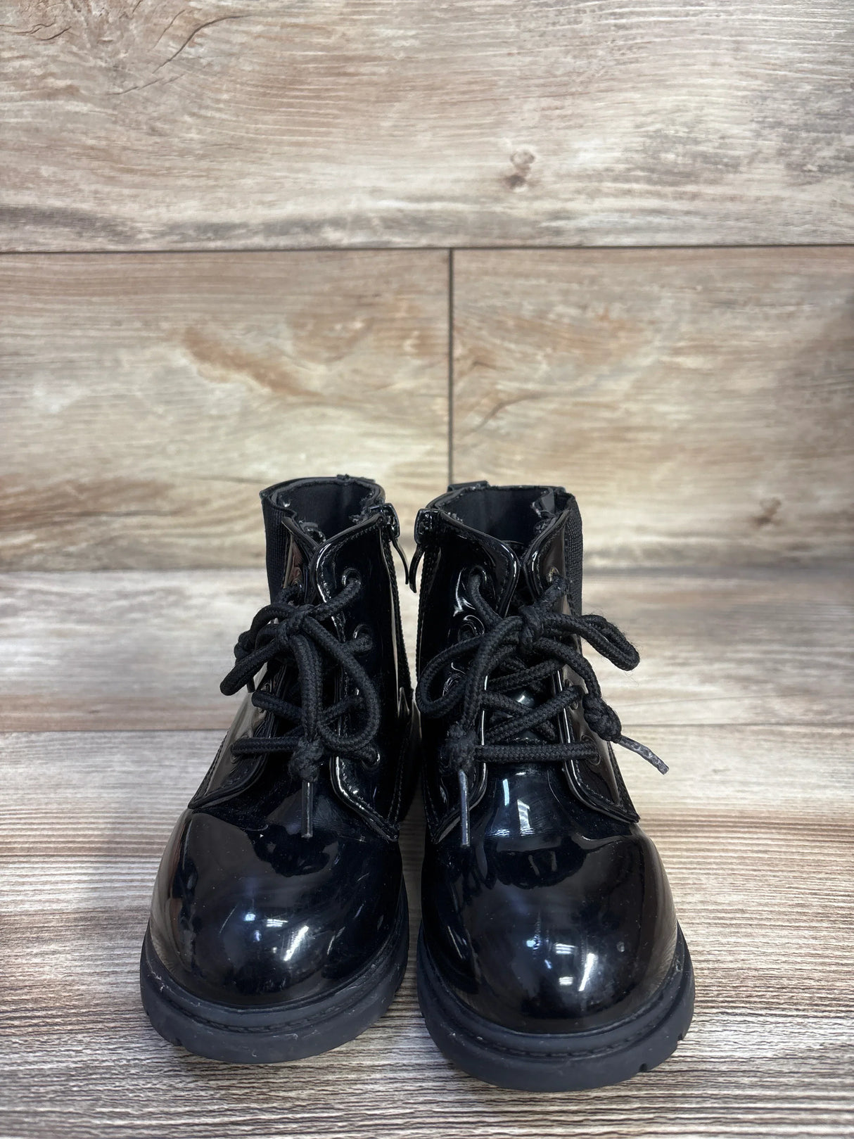 Front view of toddler black patent faux-leather lace-up ankle boots on wood floor showing round toes and laces