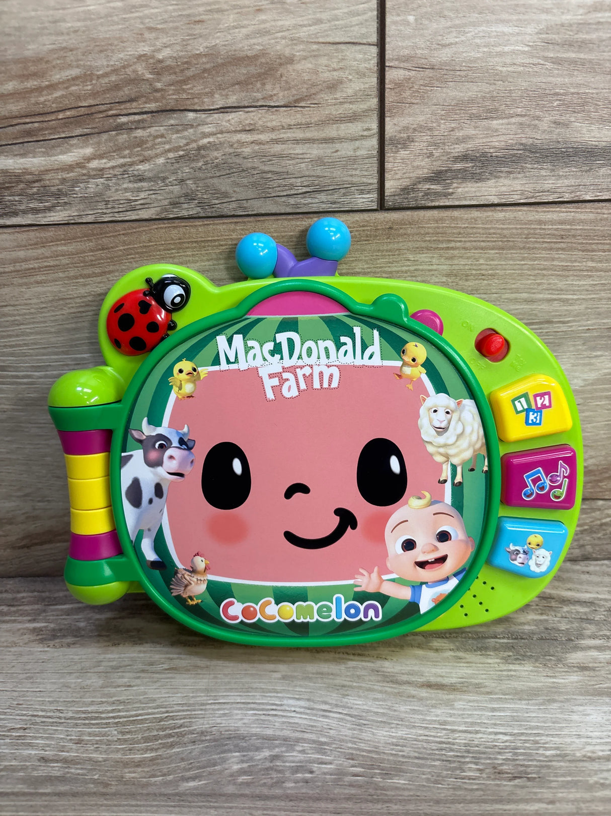 Front view of Cocomelon MacDonald Farm learning book with smiling cover, farm animals and colorful side buttons on wood floor