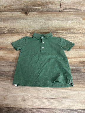 Top view of green Janie & Jack pique polo shirt size 5T showing three button placket, short sleeves and side vents on light wood background