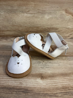 Angled front view of Little Love Bug The Sophia white baby shoes showcasing scalloped toe with heart cutout and elastic cross straps on wood background