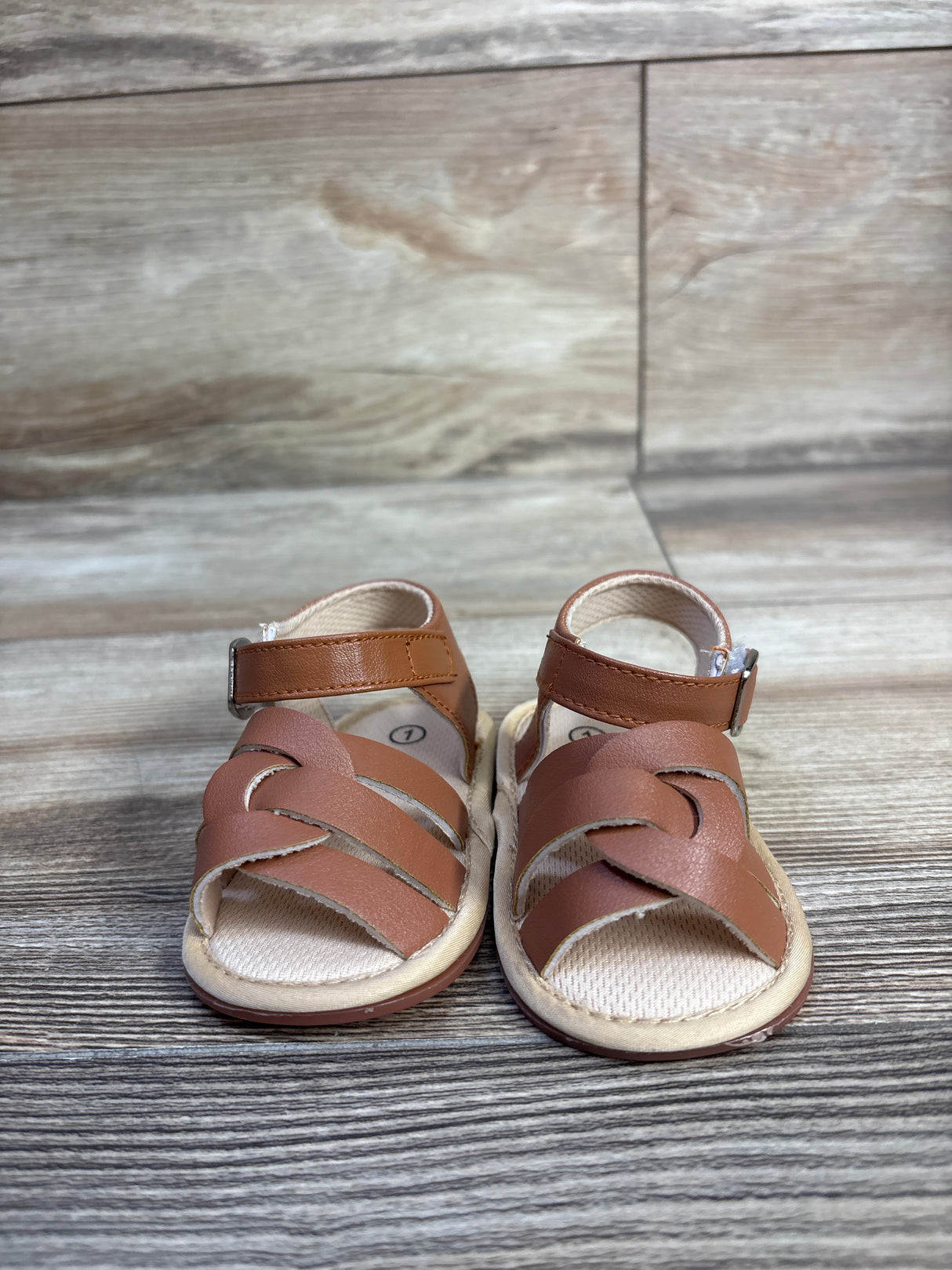 Front view of brown baby soft-sole sandals showing braided front straps, ankle strap with buckle, and cushioned insole on wood background