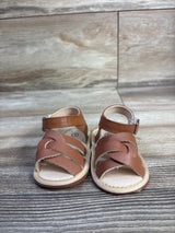 Front view of brown baby soft-sole sandals showing braided front straps, ankle strap with buckle, and cushioned insole on wood background