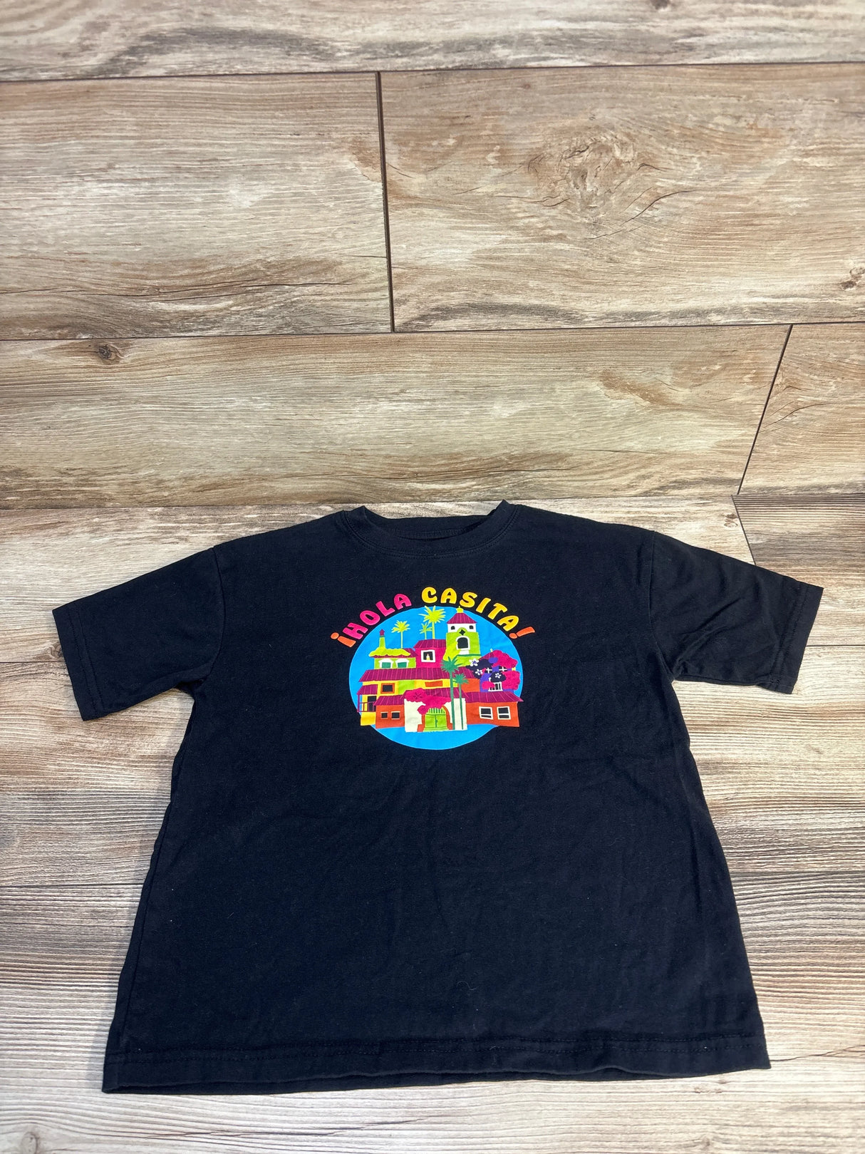 Black kids t-shirt with colorful '¡Hola Casita!' village graphic, laid on wood floor