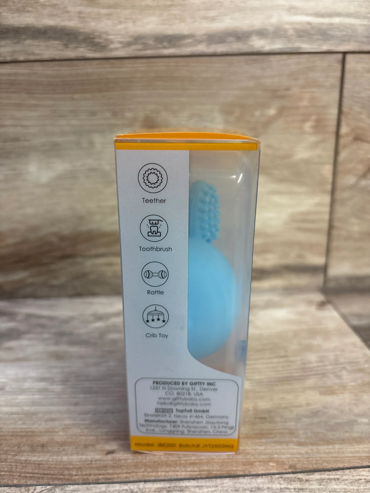 Front view of blue bunny egg teething toy in clear retail box showing textured ear nubs and icons with wood background