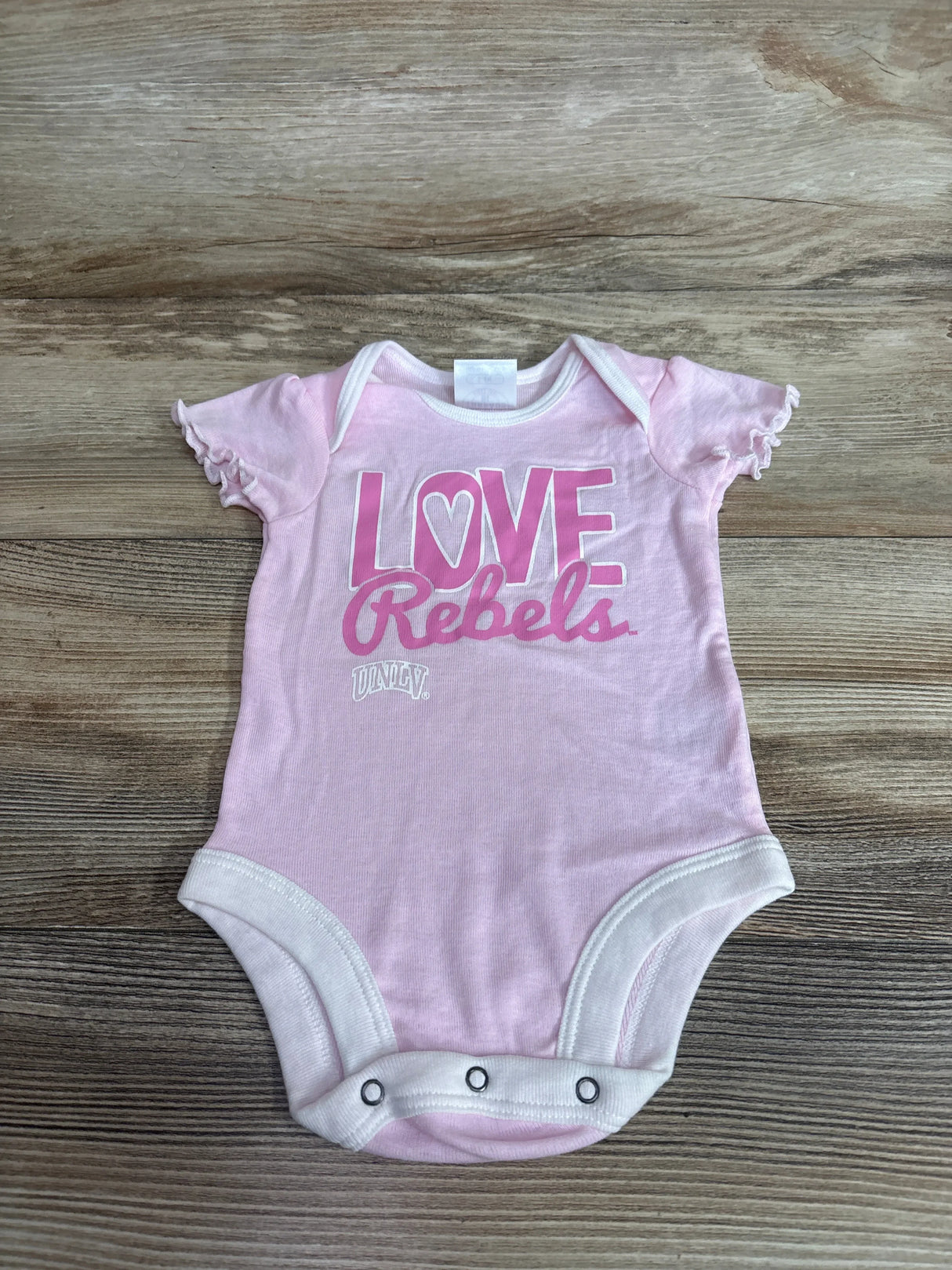 Flat lay of pink UNLV Love Rebels baby bodysuit showing ruffled cap sleeves and three snap closures on wood background