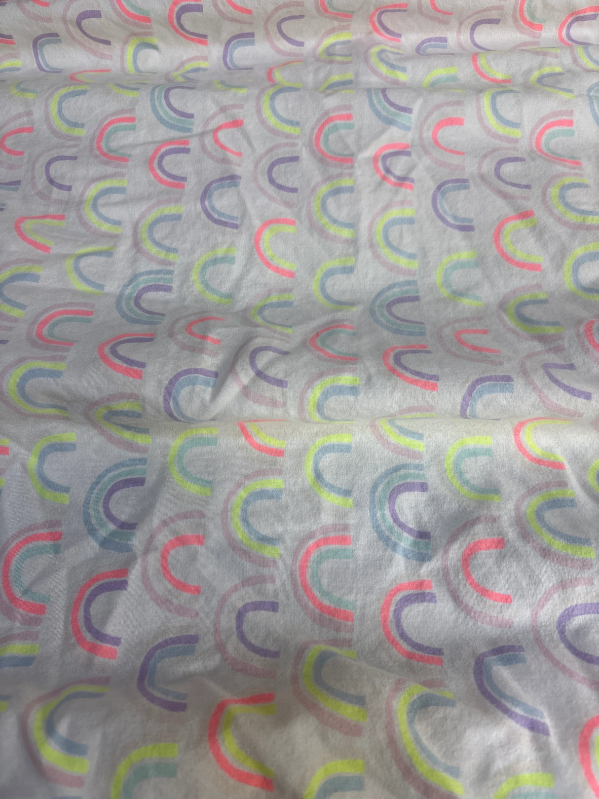 Top-down view of Cloud Island fitted crib sheet showcasing repeating pastel rainbow arc print on white background