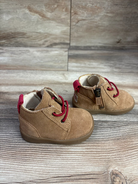 Three-quarter view of Ugg Kristjan Chukka boots with chestnut suede, red laces, and side zippers on wood background