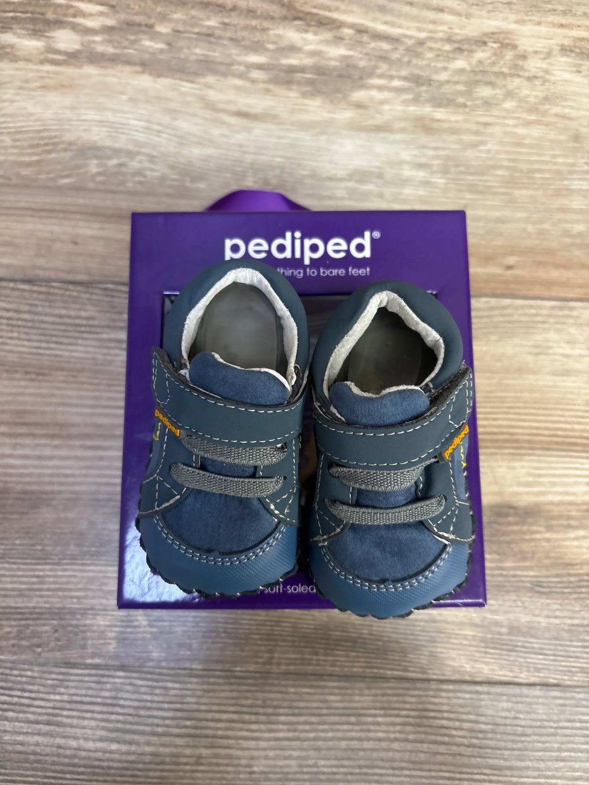 Top-down view of Pediped Dani navy crib shoes showing suede and leather upper, hook-and-loop strap and stitched outsole on a purple Pediped box
