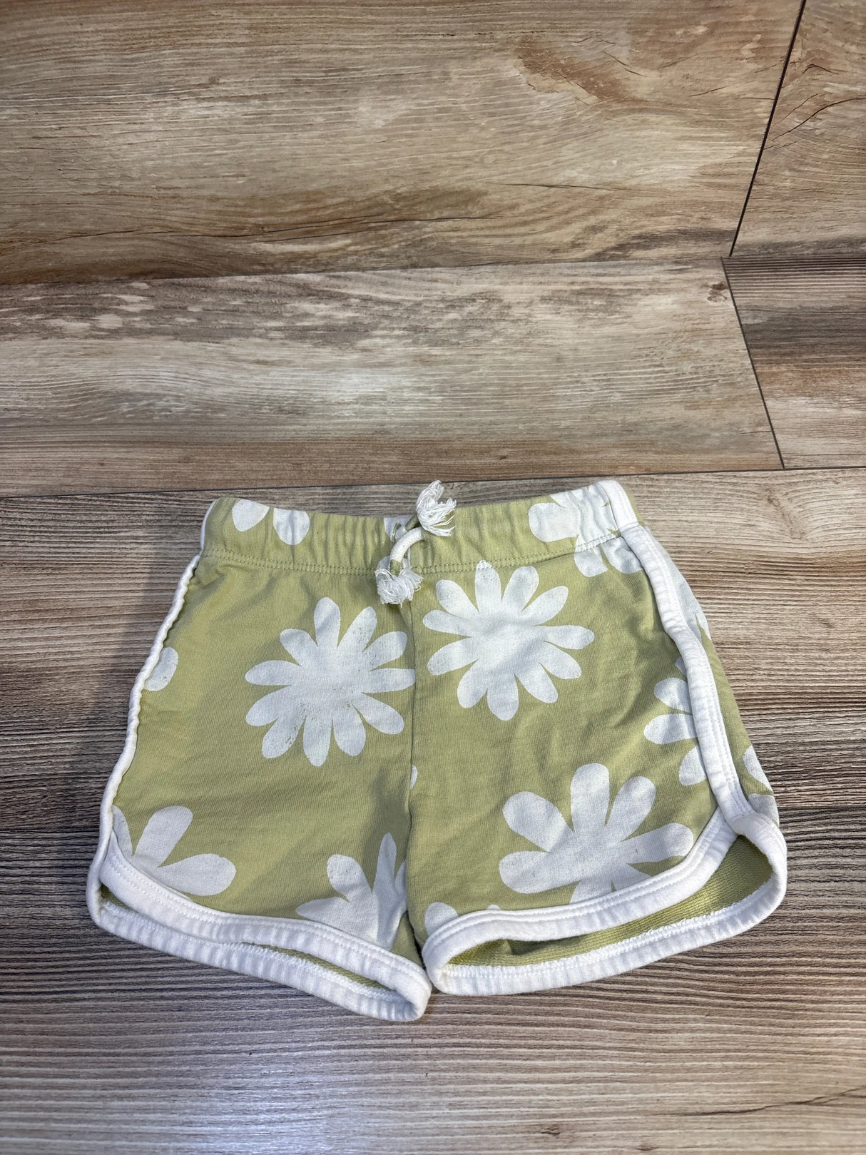 Green baby shorts with white daisy print, elastic waist, laid on wood floor