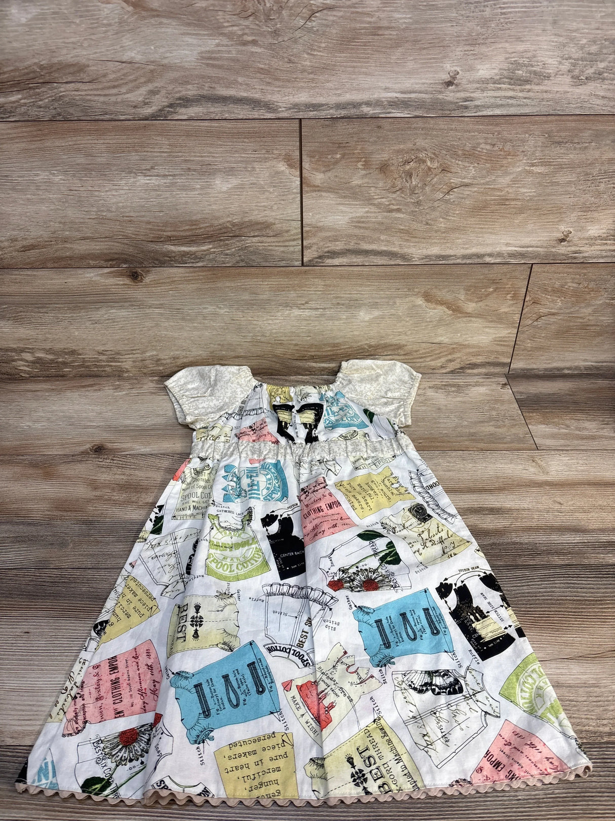 Vintage-style baby dress with sewing pattern print, ruffled sleeves, and scalloped hem on wood floor