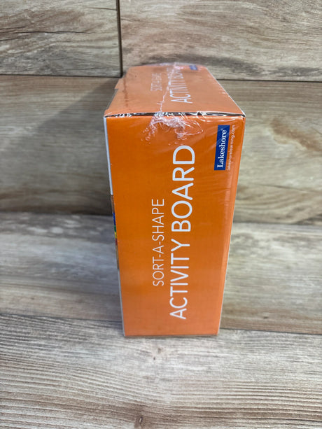 Side view of Lakeshore Sort-A-Shape Activity Board box showing orange packaging, large 'SORT-A-SHAPE ACTIVITY BOARD' text, small Lakeshore logo, and wood tabletop background